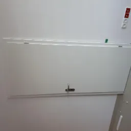 Biała klapa rewizyjna zamontowana na ścianie, widoczne gniazdka elektryczne na ścianie obok.