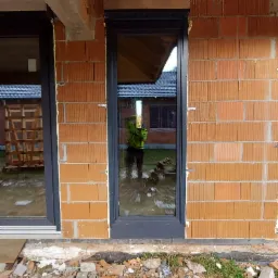 Nowe okno PCV z zamontowaną roletą zewnętrzną w surowym stanie, widoczne cegły i elementy konstrukcyjne budynku, odbicie osoby w oknie.