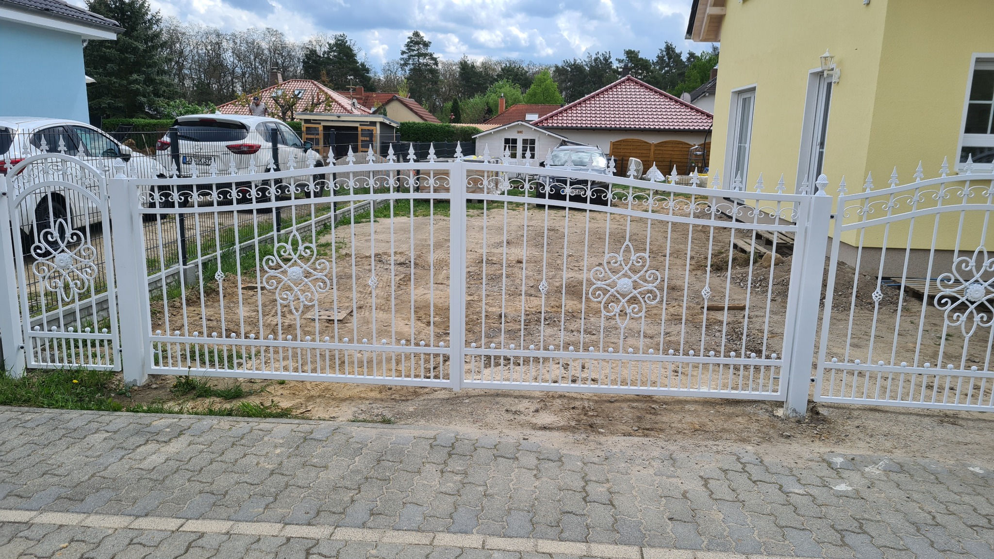 Biała brama z metalowymi zdobieniami w kształcie rozet, prowadząca na teren posesji z widocznymi samochodami i domami w tle.