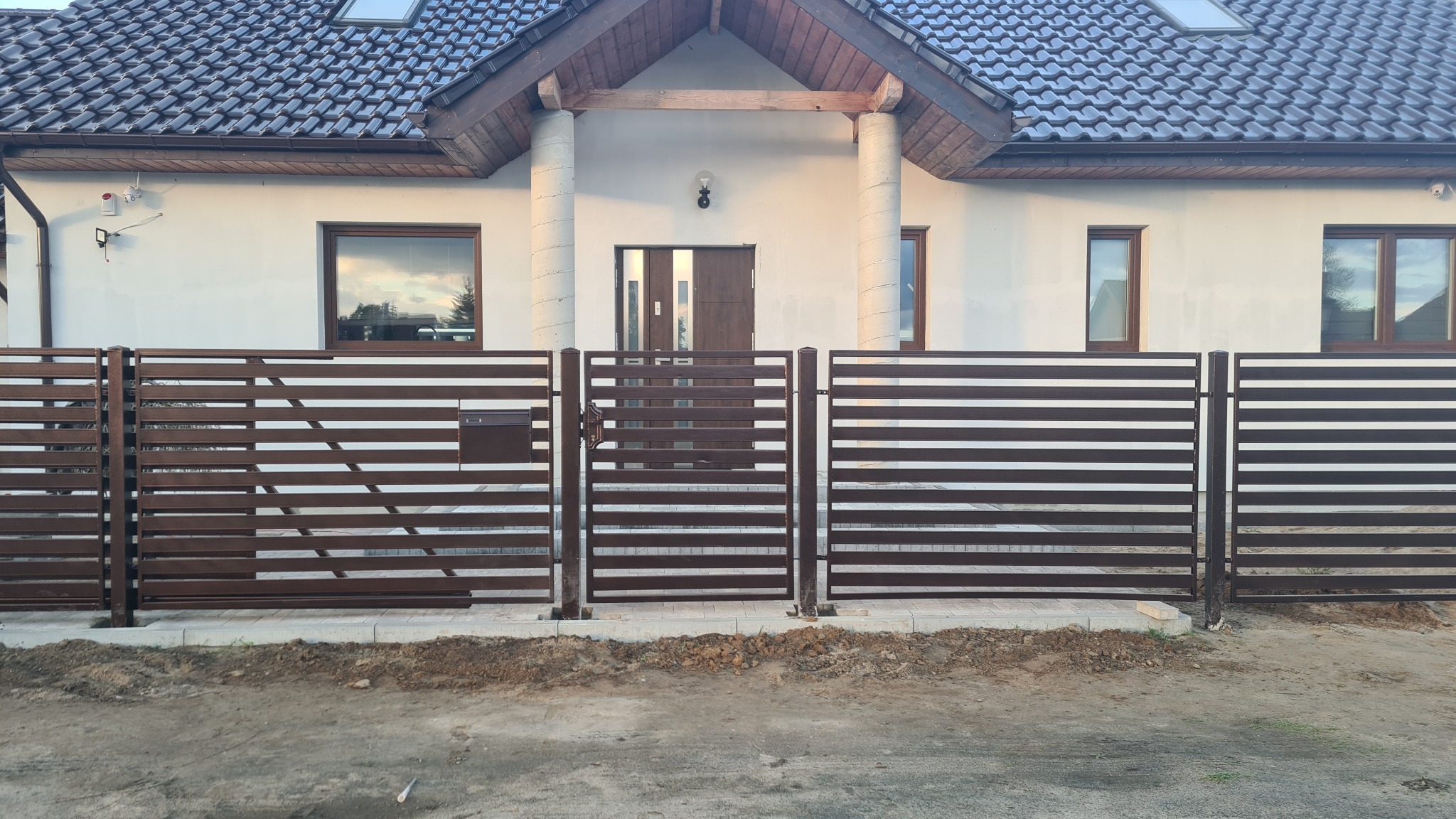 Nowoczesne brązowe ogrodzenie poziome z metalowych paneli na tle domu z białej cegły i ciemną dachówką, widoczna furtka z zamkiem i skrzynka na listy.