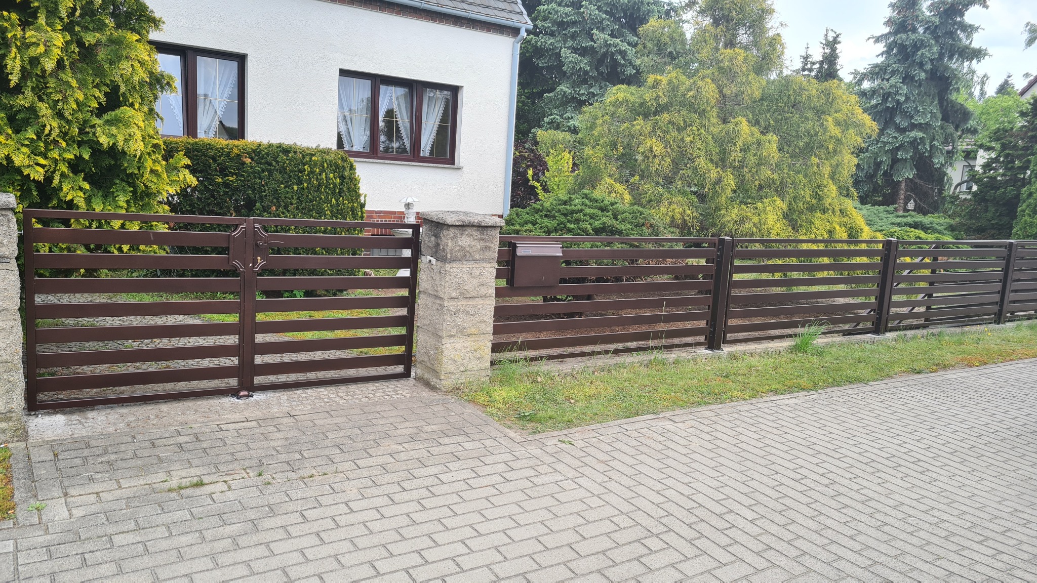 Nowoczesne, poziome brązowe przęsła ogrodzenia z metalu, brama wjazdowa z kamiennymi słupkami, skrzynka na listy w kolorze ogrodzenia, kostka brukowa na podjeździe, zieleń w tle.