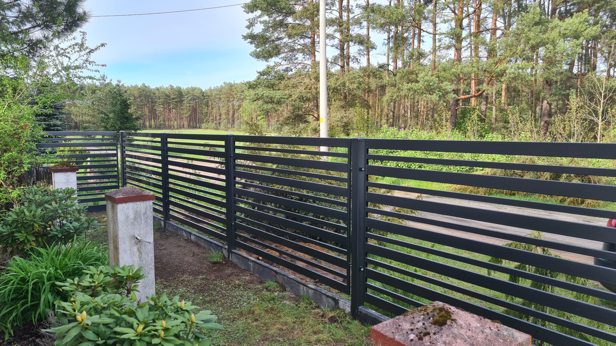 Nowoczesne, poziome, czarne ogrodzenie z metalowych profili, widok zza betonowych słupków z czerwoną nakrywką, w tle las iglasty i zielona trawa.