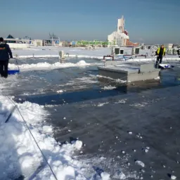 Pracownicy z łopatami odśnieżają płaski dach budynku przemysłowego, z widocznymi instalacjami i kościołem w tle, na częściowo odśnieżonej powierzchni leżą bryłki śniegu.