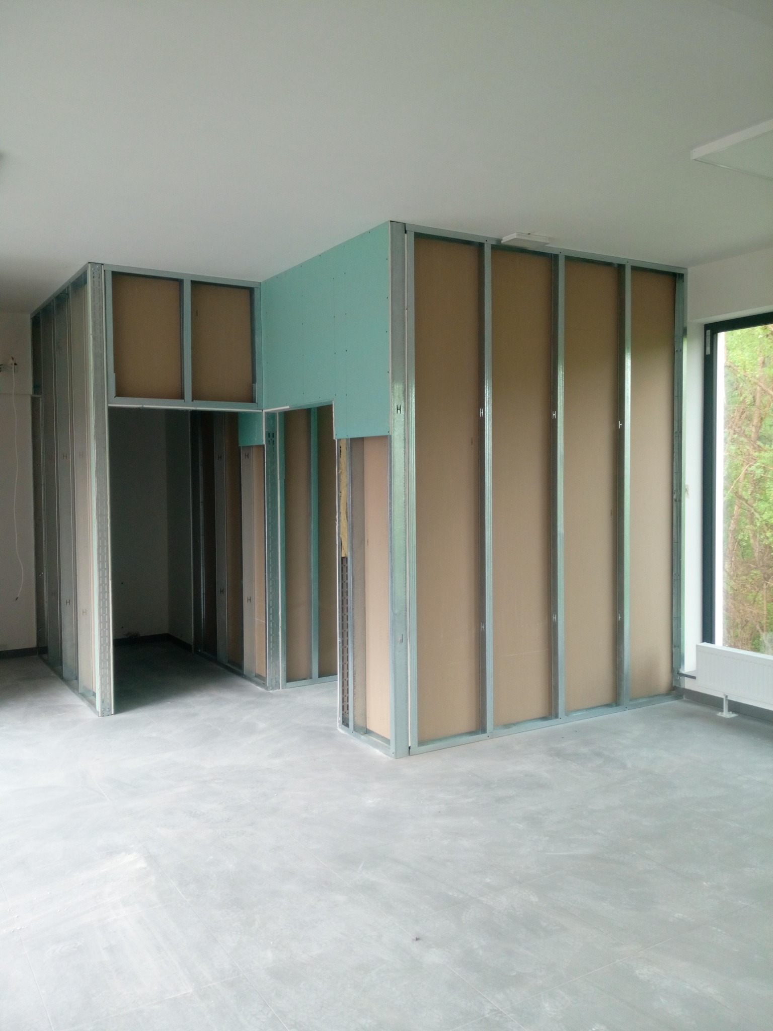 Wnętrze w trakcie montażu ścian działowych z płyt kartonowo-gipsowych na metalowym stelażu, widoczne fragmenty zielonej płyty wodoodpornej. Surowy beton na podłodze, okno z widokiem na zieleń...