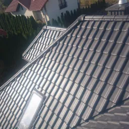 Ciemnoszara dachówka ceramiczna ułożona na dachu domu jednorodzinnego z oknem dachowym, widok z góry na tle zieleni i innych budynków.