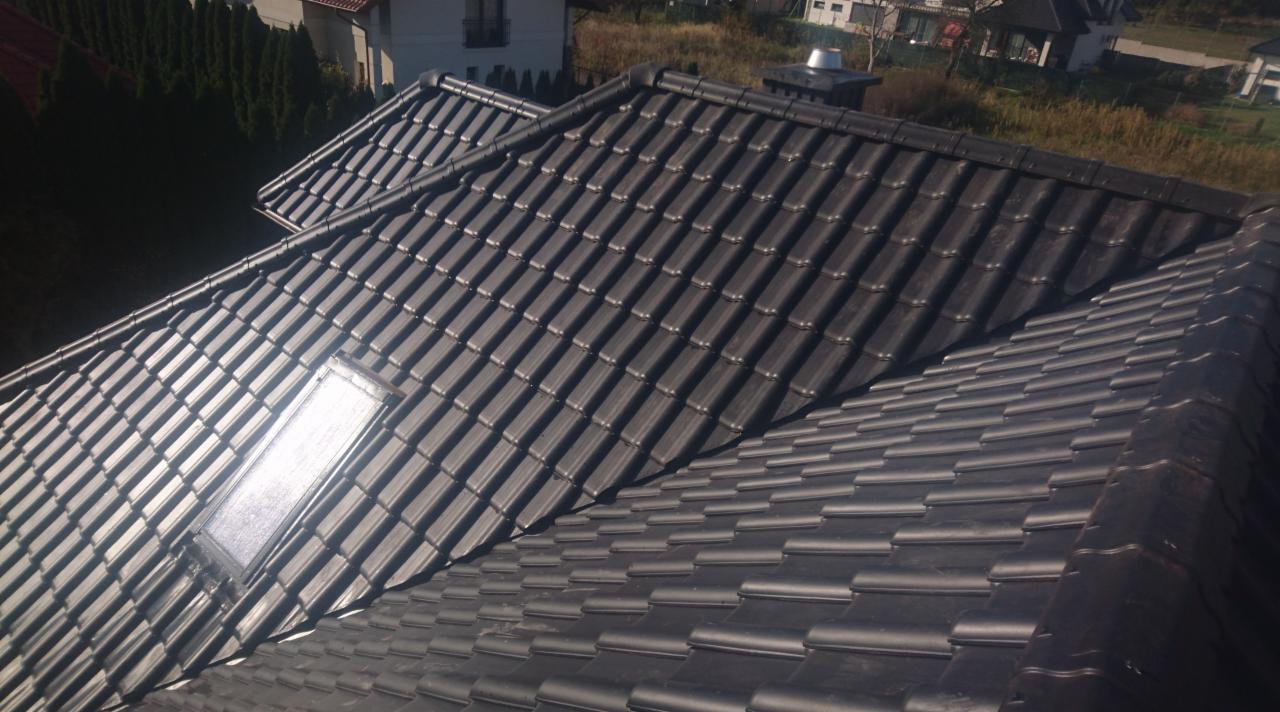 Czarna dachówka ceramiczna na dachu domu jednorodzinnego, widoczne okno dachowe odbijające światło słoneczne, w tle zabudowa mieszkalna i zieleń.
