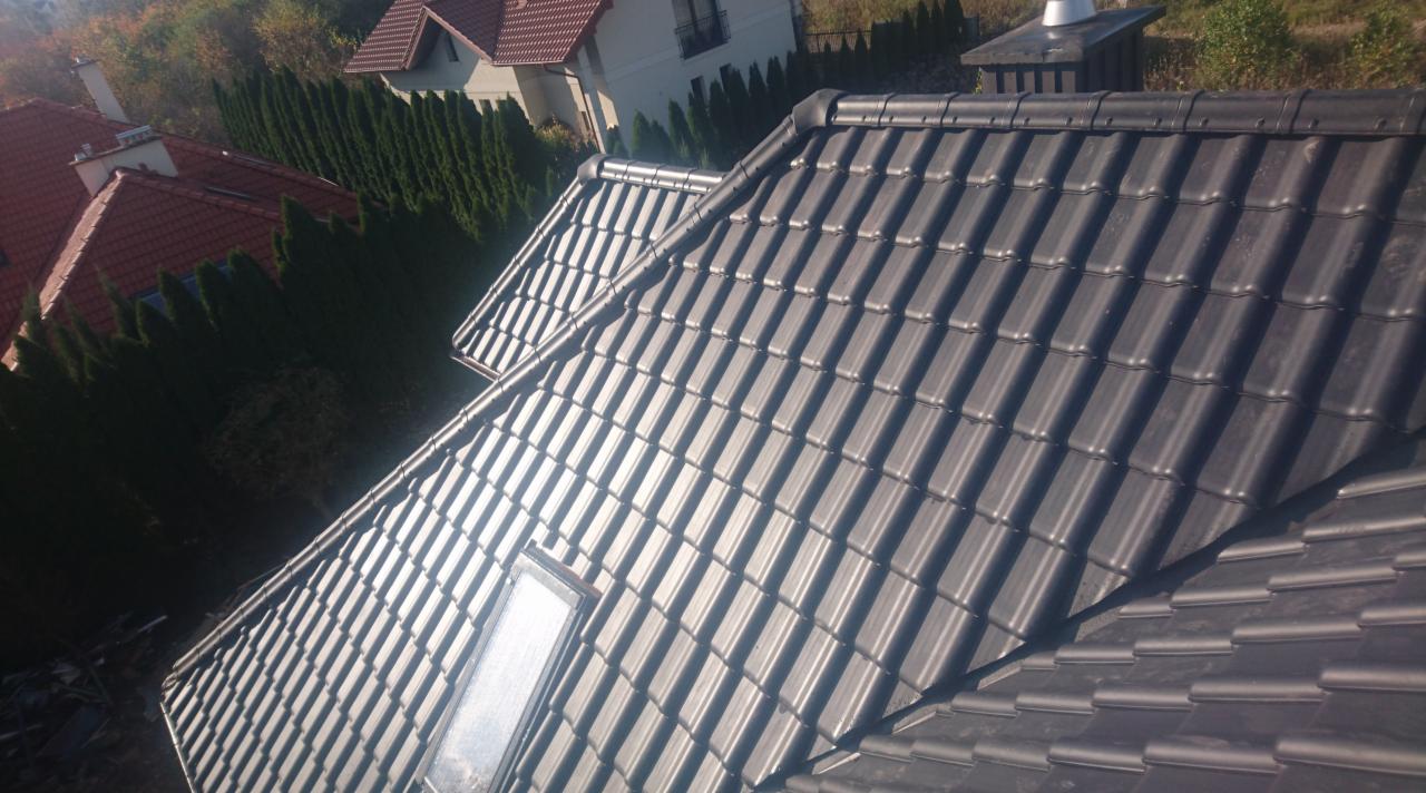 Ciemnoszara dachówka ceramiczna ułożona na dachu domu jednorodzinnego z oknem dachowym, widok z góry na tle zieleni i innych budynków.