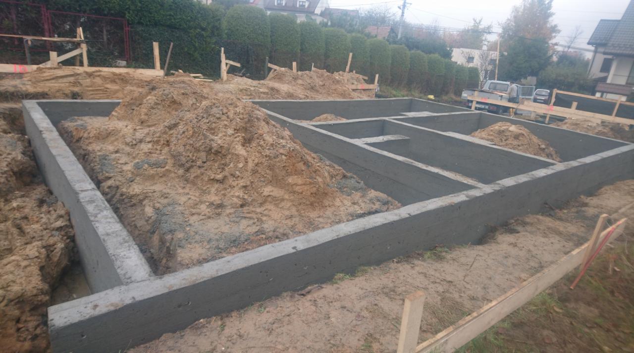 Betonowe fundamenty domu w trakcie budowy, widoczne szalunki i ziemia, w tle zabudowa jednorodzinna i krzewy.