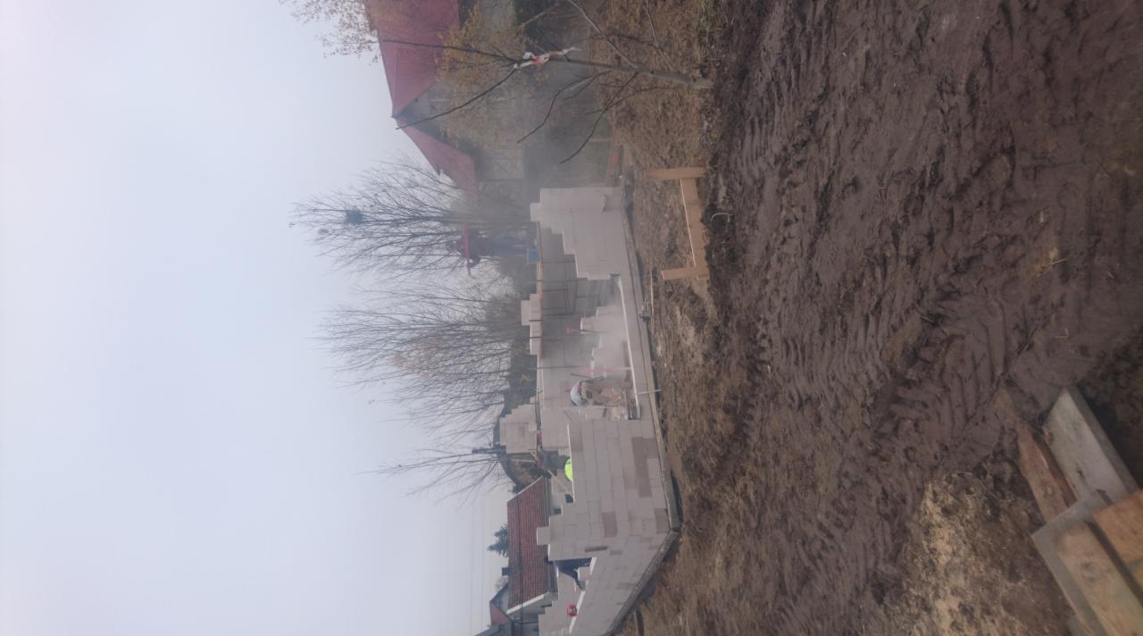 Wznoszenie ścian parterowego domu z bloczków betonu komórkowego w pochmurny dzień, widoczne ślady opon na błotnistej ziemi i fragment dachu sąsiedniego budynku.