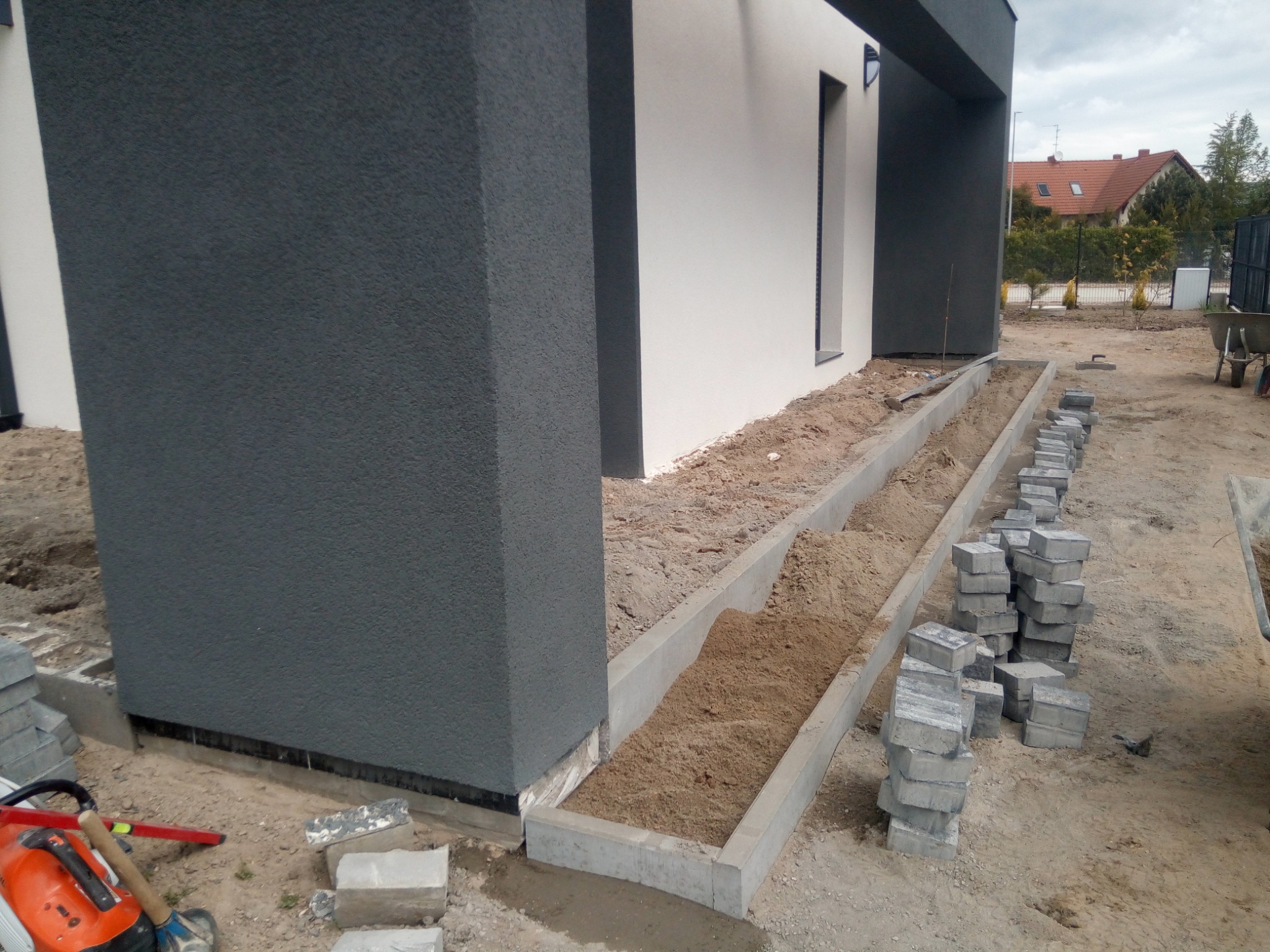 Przygotowanie podłoża pod ścieżkę z kostki brukowej przy nowoczesnym domu z szarym filarem, widoczne narzędzia i ułożone obrzeża betonowe.