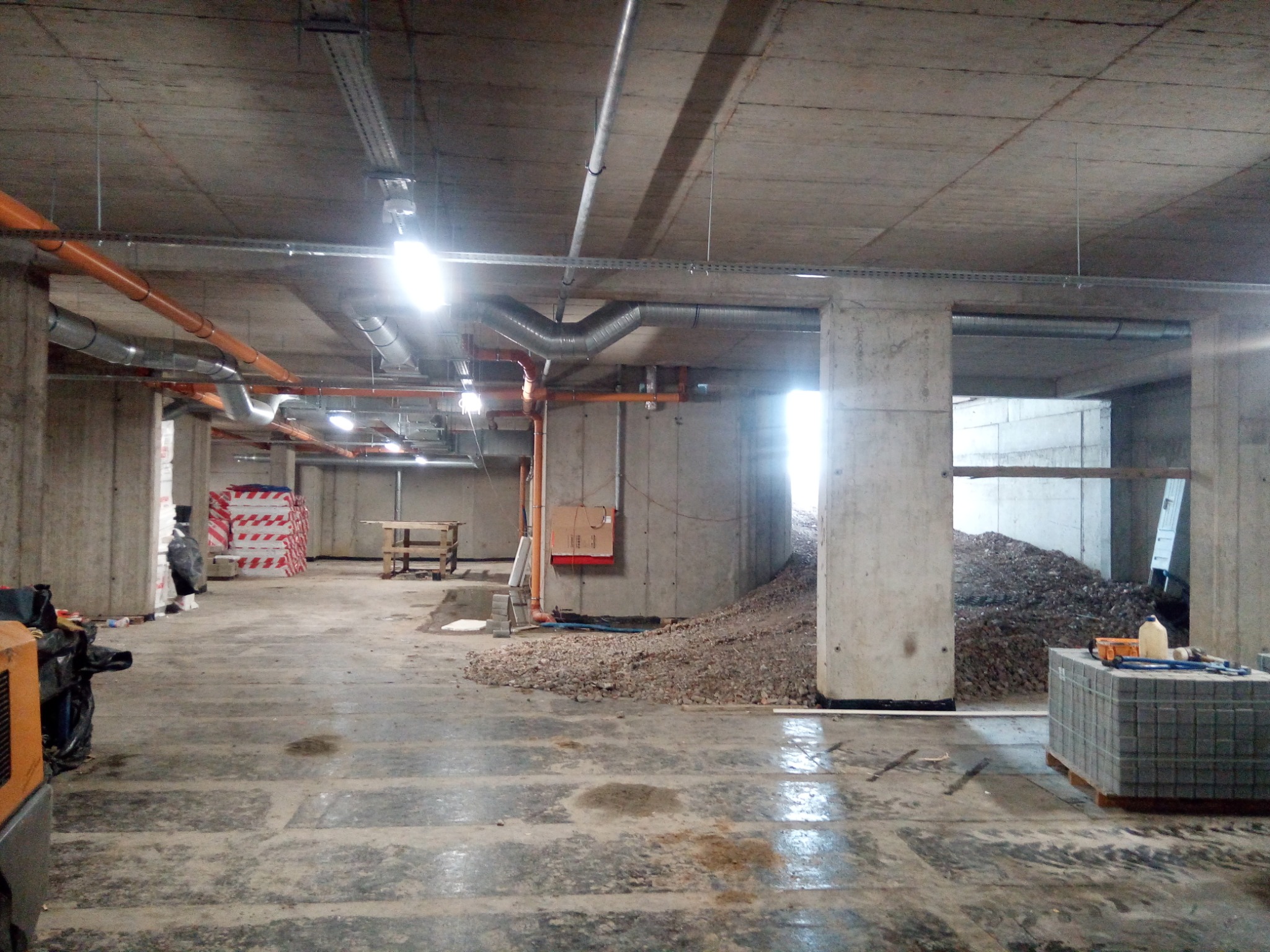 Wnętrze surowego parkingu podziemnego w trakcie budowy z betonowymi słupami, instalacjami pod sufitem, stertą gruzu i paletą kostki brukowej w tle.