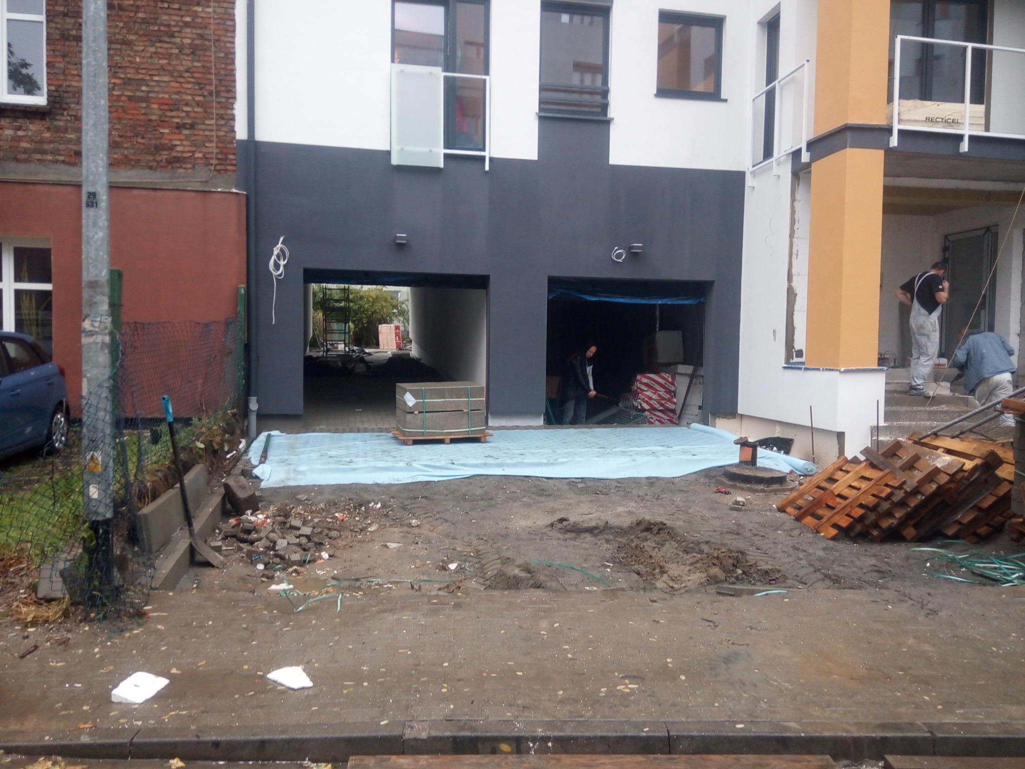 Przygotowanie terenu pod układanie kostki brukowej przed nowoczesnym budynkiem z dwoma garażami, widoczne materiały budowlane i narzędzia.