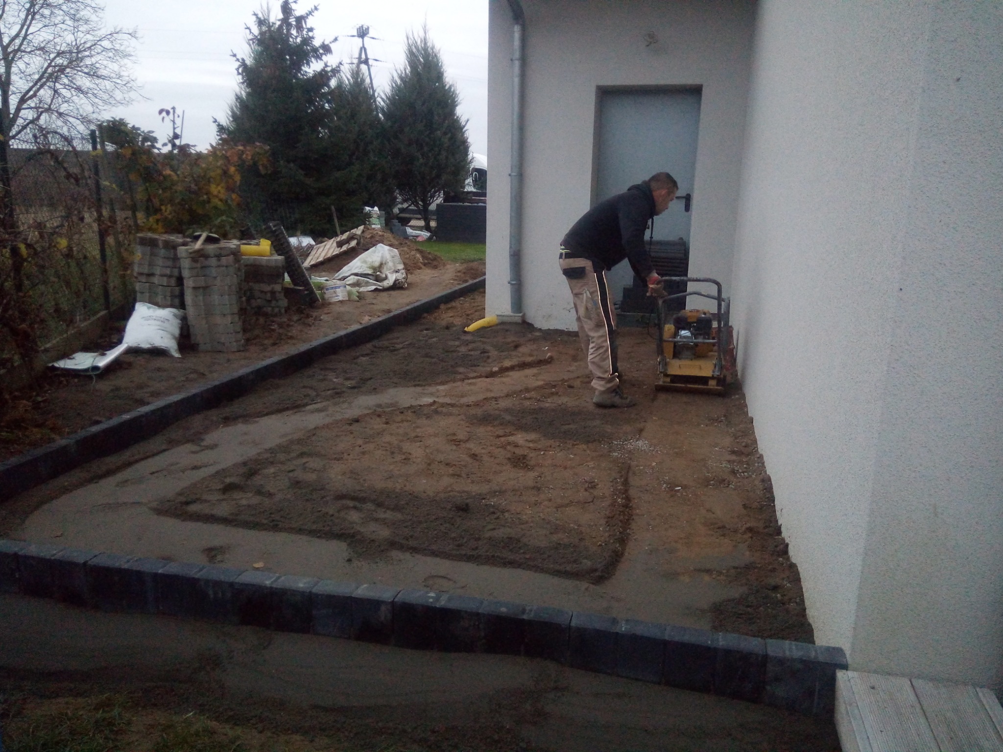 Pracownik zagęszcza podłoże pod kostkę brukową zagęszczarką wzdłuż ściany budynku, widoczne obrzeża i przygotowane kostki brukowe.