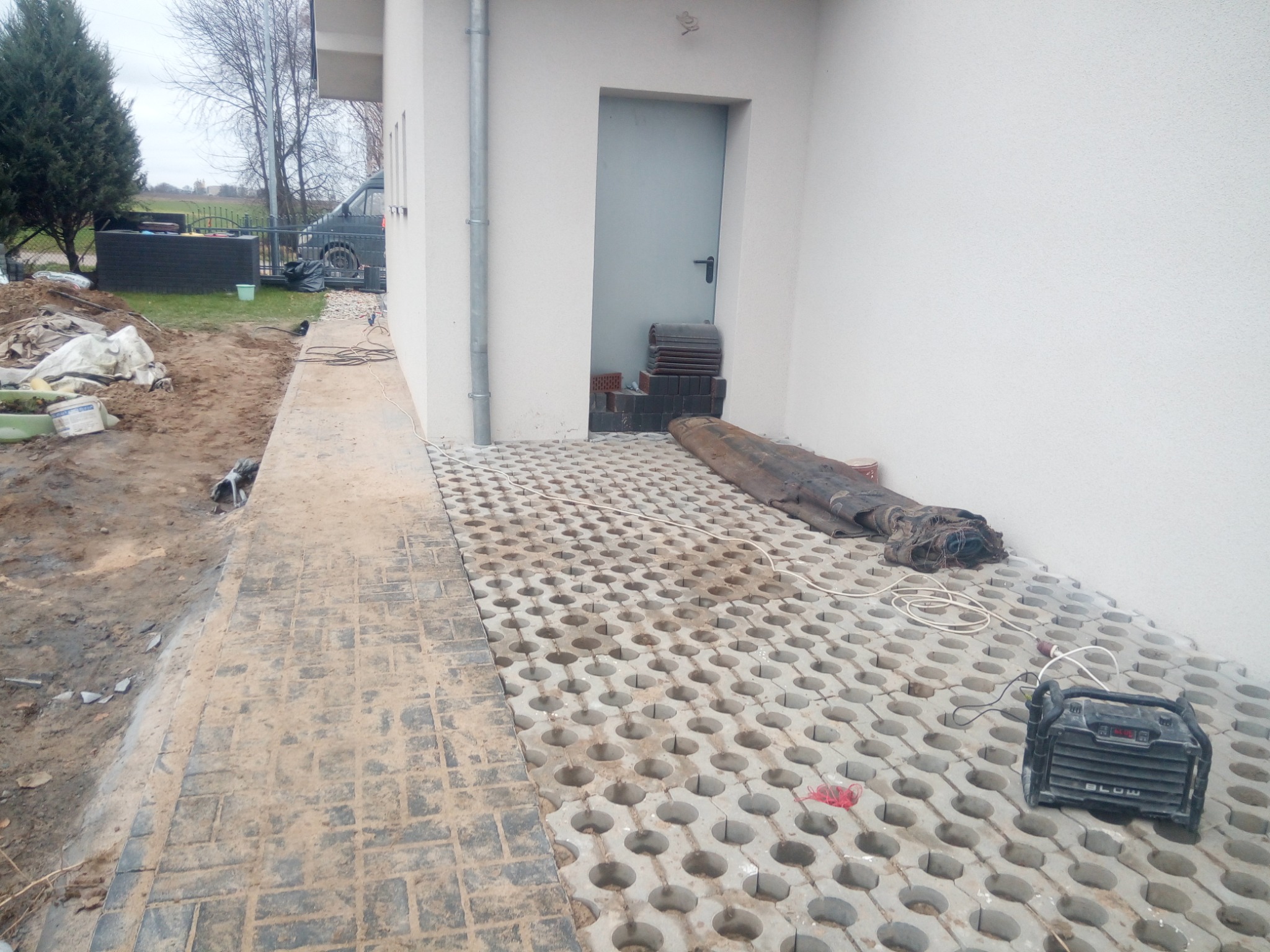 Fragment posesji z częściowo ułożoną ażurową kostką betonową, widoczne narzędzia i materiały budowlane, przygotowany teren pod dalsze prace.