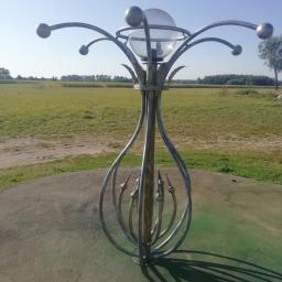 Usługi spawalnicze obrubka metali Pw-spaw - Metalowa konstrukcja z kulami i szklaną sferą na szczycie, przypominająca lampę, umieszczona na betonowym postumencie na tle zielonego pola i lasu.