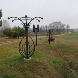 Usługi spawalnicze obrubka metali Pw-spaw - Ozdobne, metalowe konstrukcje w kształcie kwiatów i geometrycznych form na tle wiejskiego krajobrazu z polną drogą w pochmurny dzień.