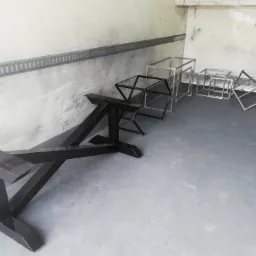 Metalowe konstrukcje w surowym wnętrzu, czarna podstawa stołu na pierwszym planie, za nią srebrne ramy w kształcie sześcian&oacute;w i tr&oacute;jkąt&oacute;w, widoczne ślady po spawaniu.