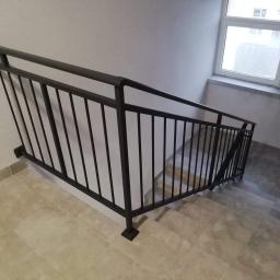 Usługi spawalnicze obrubka metali Pw-spaw - Metalowa poręcz schodowa w ciemnym kolorze, zamontowana wewnątrz klatki schodowej, widok z góry na schody wyłożone płytkami.