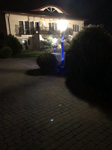 Nocne ujęcie dwupiętrowego domu z balkonami zabezpieczonymi metalowymi barierkami, oświetlonego przez lampę uliczną z niebieskim podświetleniem, widoczne z brukowanej ścieżki.