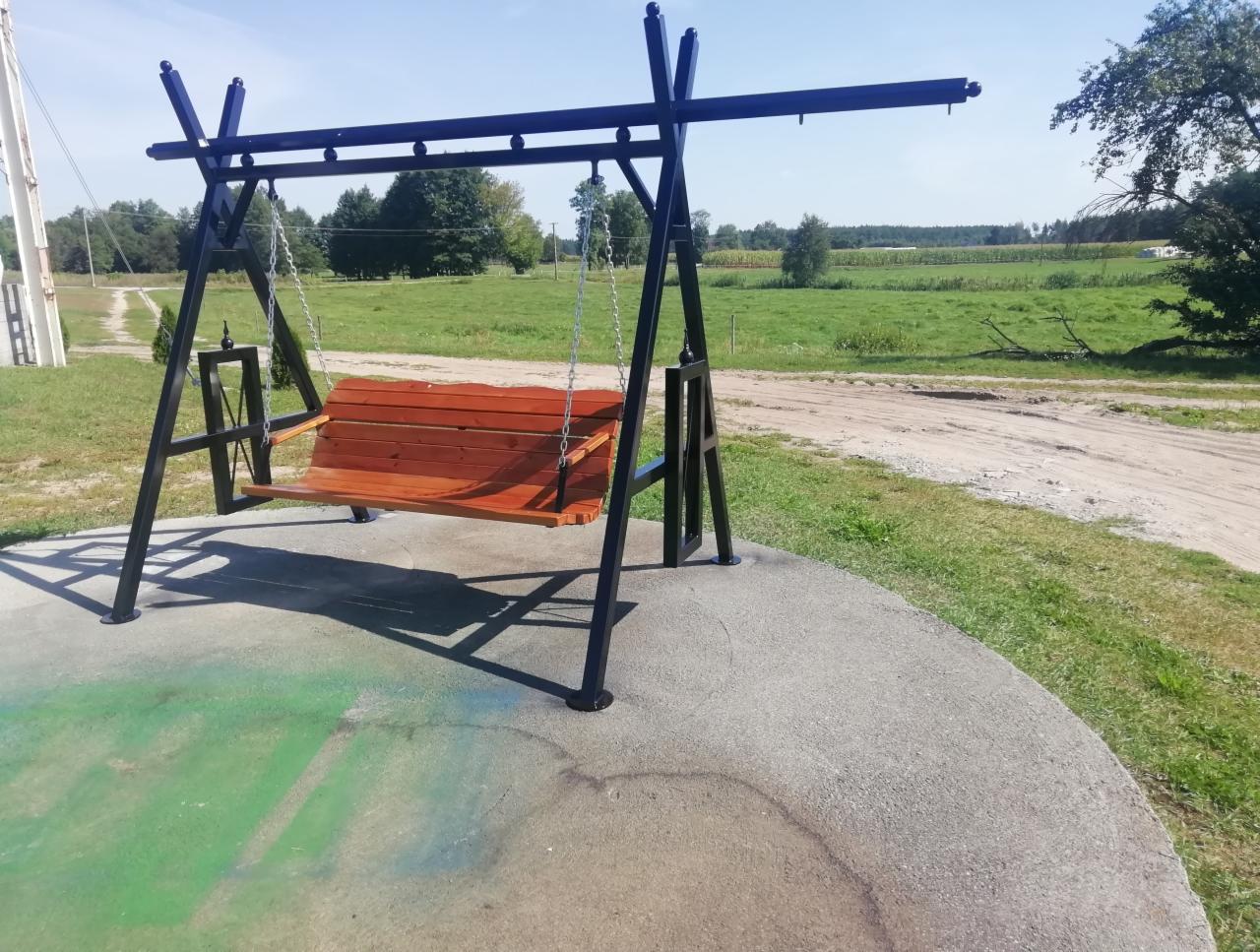 Metalowa konstrukcja huśtawki z drewnianym siedziskiem, łańcuchami i ozdobnymi zakończeniami, umieszczona na betonowym podłożu z zielonymi akcentami, w tle krajobraz wiejski.