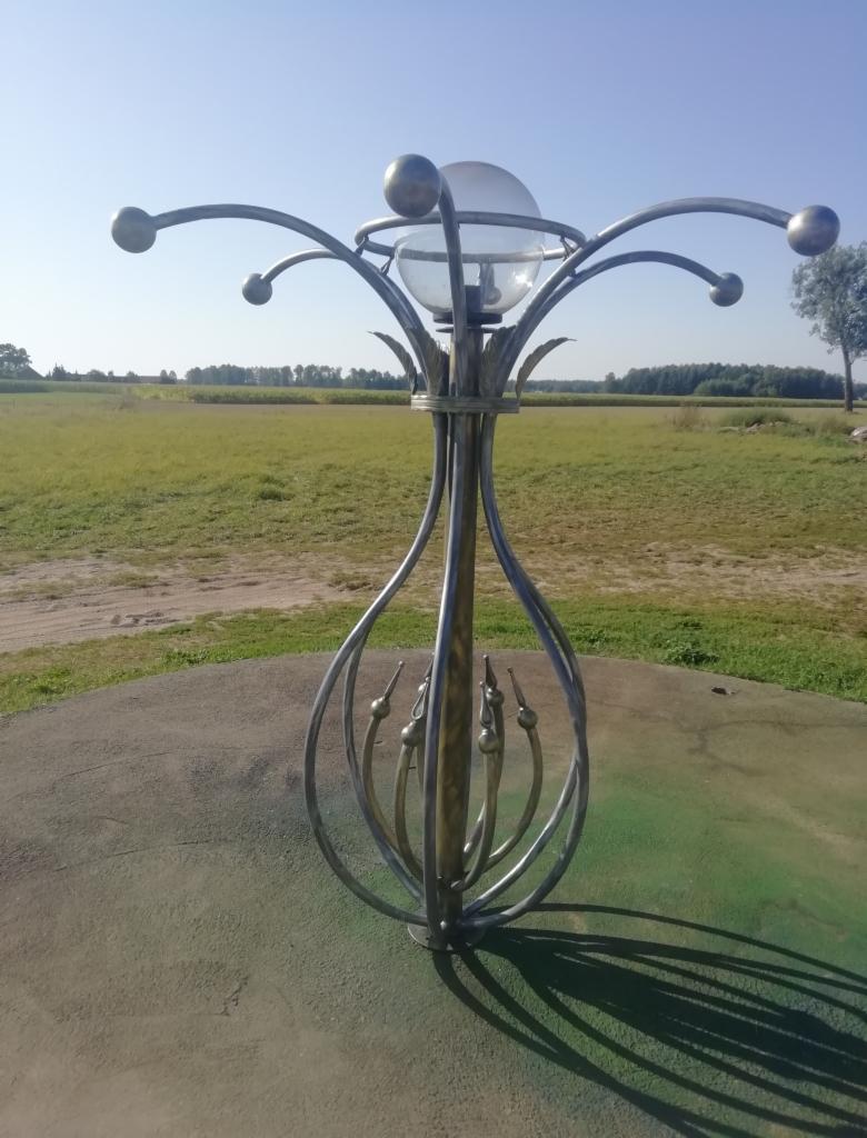 Metalowa konstrukcja z kulami i szklaną sferą na szczycie, przypominająca lampę, umieszczona na betonowym postumencie na tle zielonego pola i lasu.