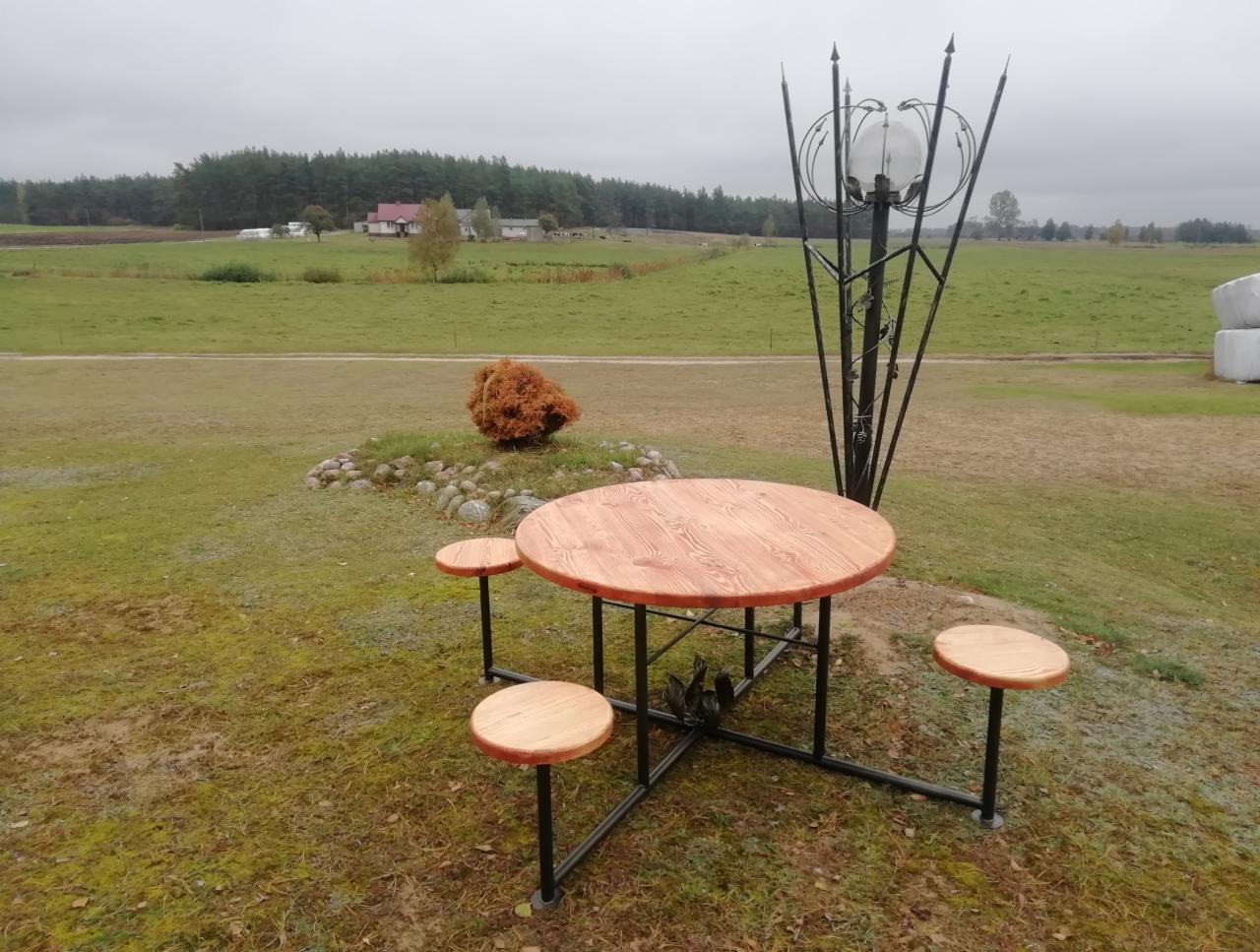 Okrągły drewniany stół z trzema taboretami na czarnej, metalowej konstrukcji, obok ozdobna, metalowa lampa z kulistym kloszem, w tle rozległy krajobraz polny i zabudowania gospodarcze.