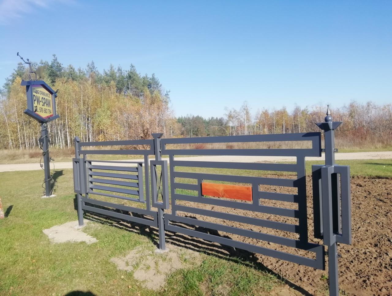 Nowoczesna, szara brama metalowa z drewnianym akcentem, widoczna tablica reklamowa firmy na tle jesiennego krajobrazu.