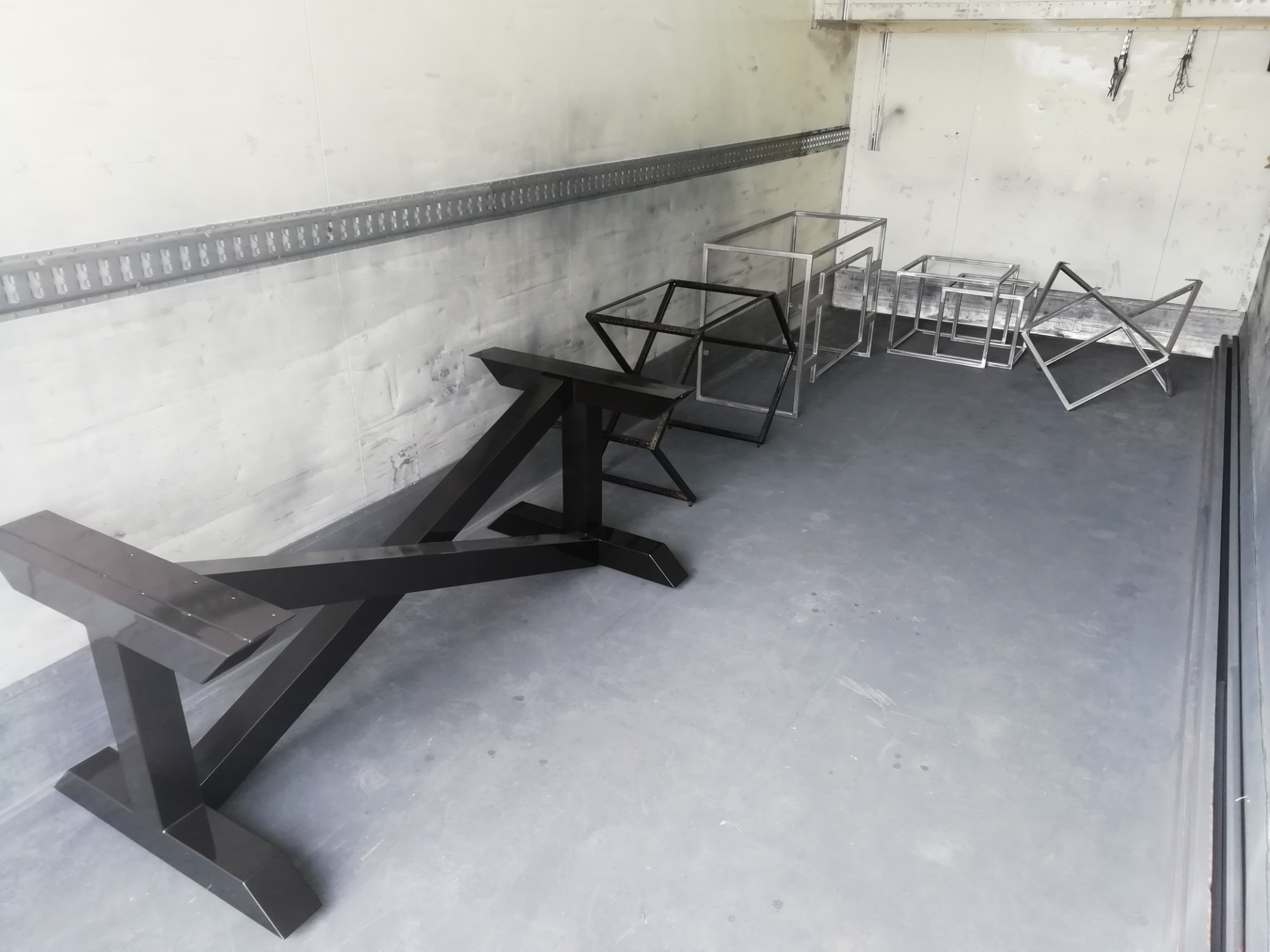 Metalowe konstrukcje w surowym wnętrzu, czarna podstawa stołu na pierwszym planie, za nią srebrne ramy w kształcie sześcianów i trójkątów, widoczne ślady po spawaniu.