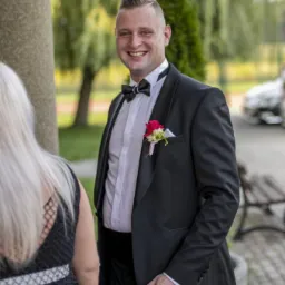 Uśmiechnięty mężczyzna w czarnym smokingu z butonierką, stojący obok kobiety z blond włosami, na tle drzew i auta. Ujęcie plenerowe.