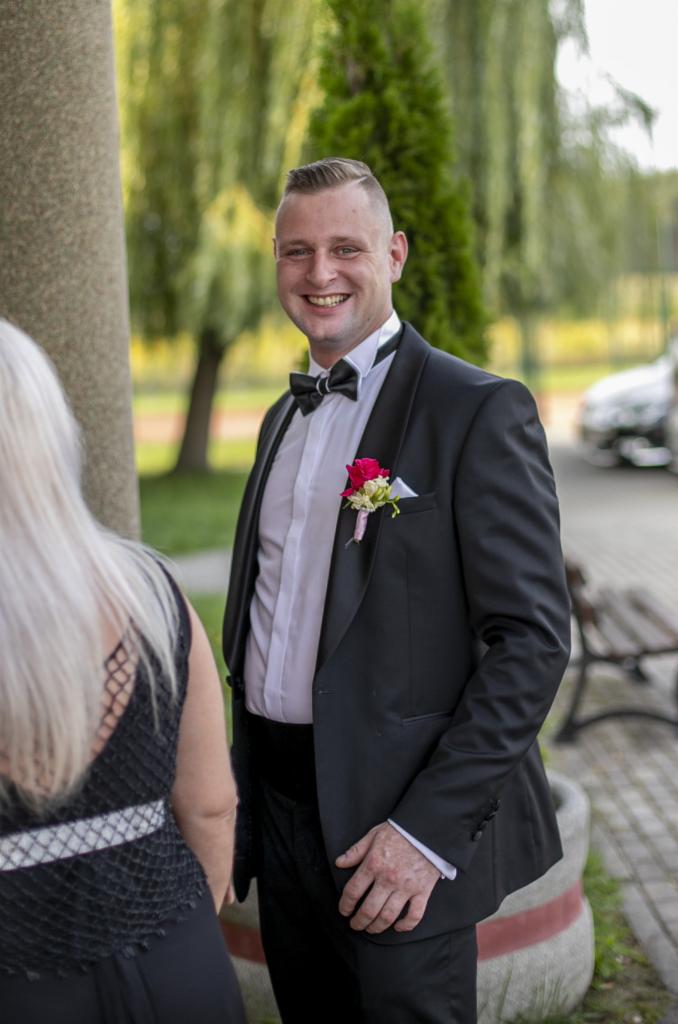 Uśmiechnięty mężczyzna w czarnym smokingu z butonierką, stojący obok kobiety z blond włosami, na tle drzew i auta. Ujęcie plenerowe.