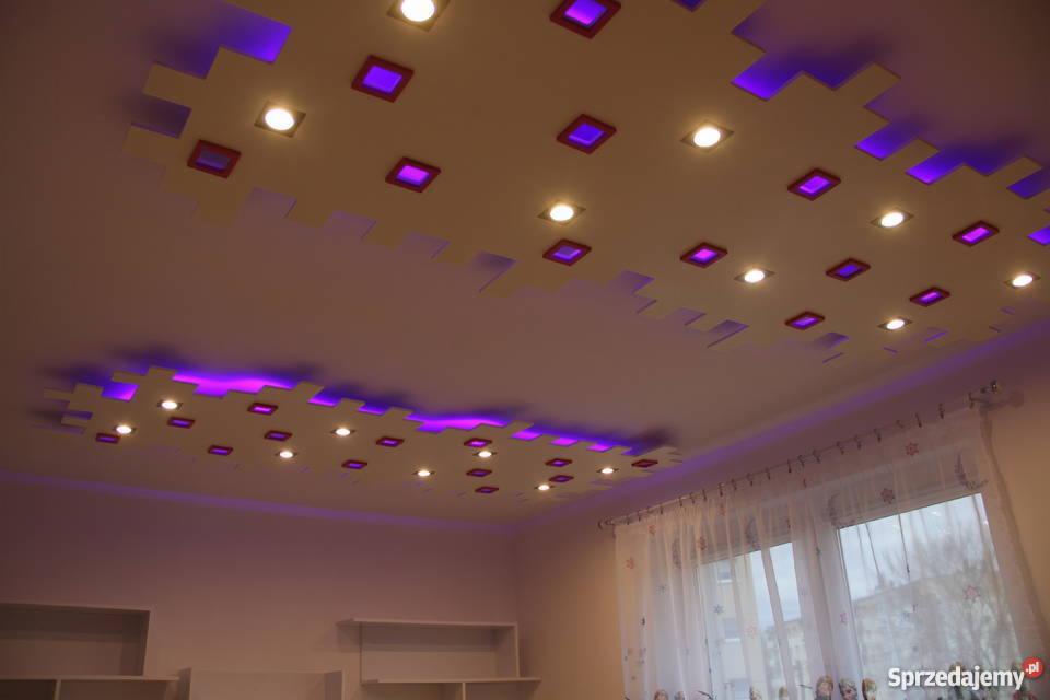Sufit podwieszany z nieregularnymi wgłębieniami, podświetlony fioletowym światłem LED, z wbudowanymi punktowymi lampami halogenowymi, widok z dołu na tle okna z firanką.