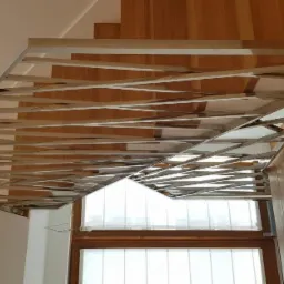 Nowoczesna balustrada ze stali nierdzewnej z poziomymi, krzyżującymi się elementami, zamontowana przy drewnianych schodach, widok od dołu w kierunku okna.