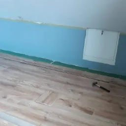 Fragment układania jasnych paneli podłogowych w pokoju ze skośnym sufitem i niebieskimi ścianami; widoczny młotek i niedopasowany panel.