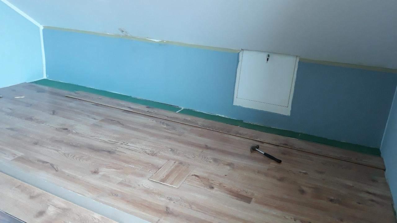 Fragment układania jasnych paneli podłogowych w pokoju ze skośnym sufitem i niebieskimi ścianami; widoczny młotek i niedopasowany panel.