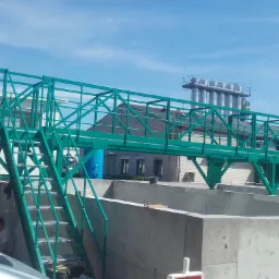 Zielona, metalowa konstrukcja pomostowa z kratowymi schodami, prawdopodobnie na terenie oczyszczalni ścieków, widoczny fragment samochodu dostawczego i pracownik w cieniu.