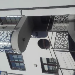 Nowoczesny budynek z balkonem i ażurowymi elementami zabezpieczającymi, widoczne okrągłe okno i alarm na ścianie.