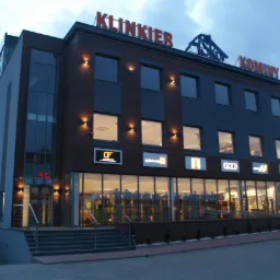 Fasada budynku z cegły klinkierowej z oknami i oświetleniem zewnętrznym, na dachu widoczne napisy 'KLINKIER' i 'KOMINY', a nad wejściem szyldy reklamowe różnych firm.