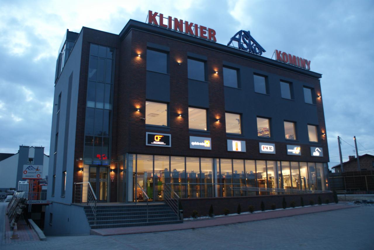 Fasada budynku z cegły klinkierowej z oknami i oświetleniem zewnętrznym, na dachu widoczne napisy 'KLINKIER' i 'KOMINY', a nad wejściem szyldy reklamowe różnych firm.