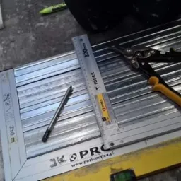 Przygotowanie profili metalowych do montażu, widoczne narzędzia: nożyce do blachy z żółtymi uchwytami, ołówek, miarka Topex, ułożone na ramie z profili.