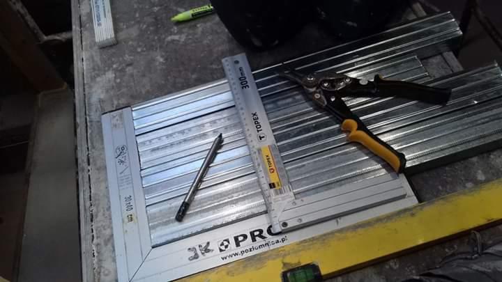 Przygotowanie profili metalowych do montażu, widoczne narzędzia: nożyce do blachy z żółtymi uchwytami, ołówek, miarka Topex, ułożone na ramie z profili.