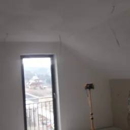 Pokój na poddaszu po nałożeniu gładzi, zabezpieczony folią malarską, z widocznymi przewodami i lampą na statywie.