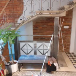 Klatka schodowa z ceglaną ścianą, dekoracyjną balustradą i wzorzystą posadzką, uzupełniona roślinami doniczkowymi i ławką z poduszką.