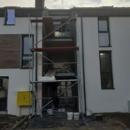 Budynek w trakcie prac elewacyjnych, widoczne rusztowanie, puszki z farbą, zabezpieczona folią podstawa budynku, fragment ocieplonego fragmentu ściany, oraz osoba pracująca na rusztowaniu.