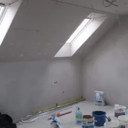 Poddasze w trakcie wykańczania: zamontowane płyty kartonowo-gipsowe na skosach dachu z oknami, częściowo wygładzone ściany, widoczne materiały budowlane i narzędzia przygotowane do dalszych prac.