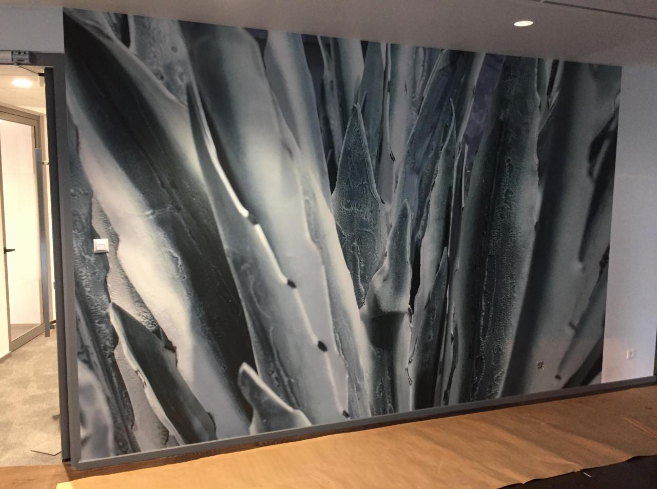 Ściana pokryta fototapetą z abstrakcyjnym wzorem przypominającym liście w odcieniach szarości i bieli, widoczny fragment podłogi zabezpieczony folią malarską, drzwi wejściowe w tle.