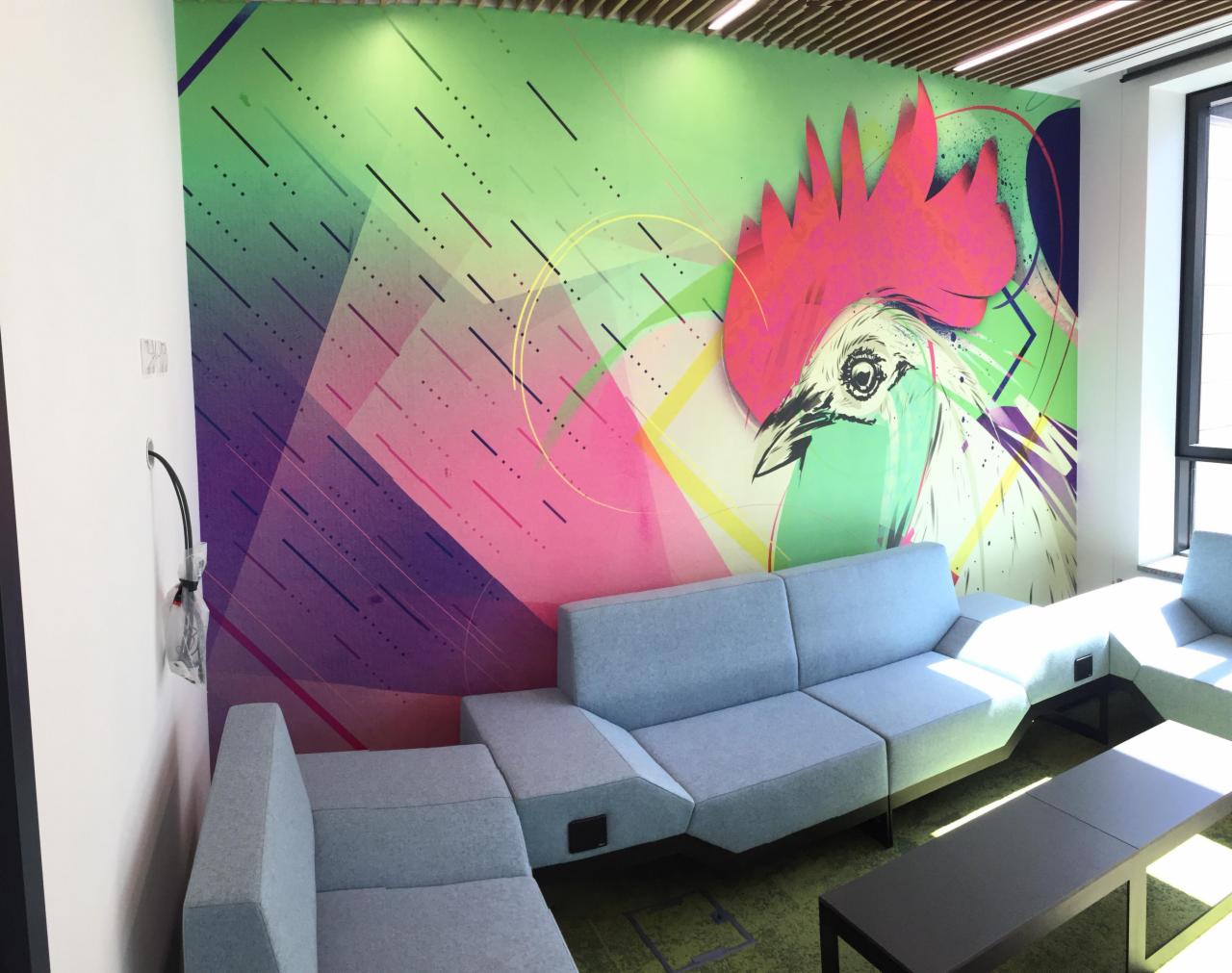 Wnętrze biurowe z fototapetą przedstawiającą abstrakcyjnego koguta na ścianie, uzupełnione modułową sofą w odcieniach szarości.