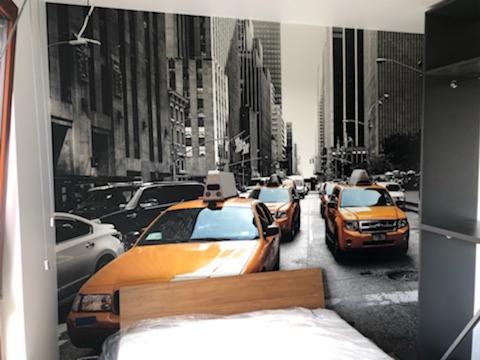 Tapeta w sypialni przedstawiająca czarno-biały widok ulicy w Nowym Jorku z żółtymi taksówkami, naklejona na całą ścianę za łóżkiem.