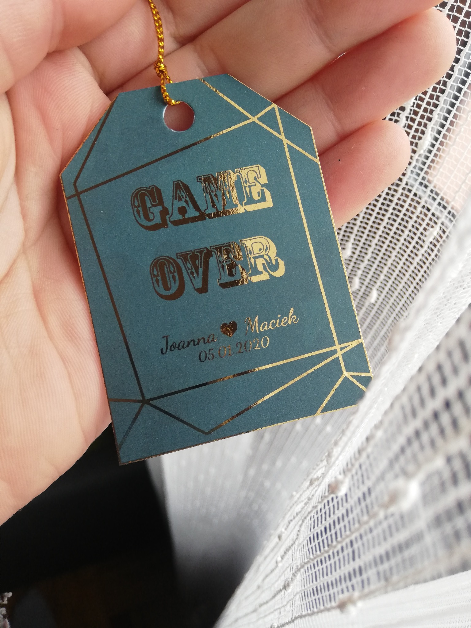 Turkusowa zawieszka z napisem 'GAME OVER', imionami Joanna i Maciek oraz datą 05.01.2020, zdobiona złotymi geometrycznymi wzorami i zawieszona na złotym sznurku, trzymana w dłoni na tle białej...