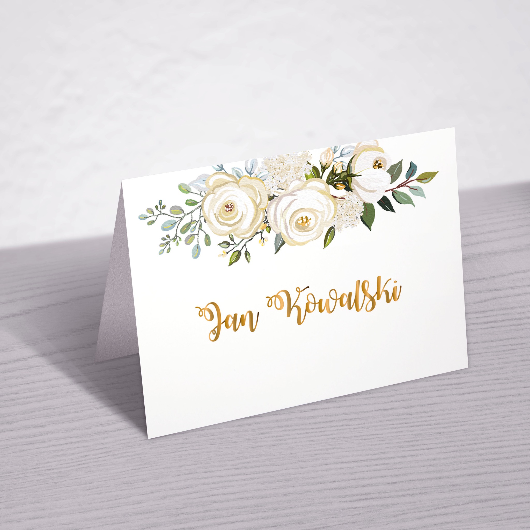 Elegancki, składany wizytownik z białym tłem, zdobiony akwarelowymi białymi różami i złotym napisem 'Jan Kowalski', leżący na szarej, drewnianej powierzchni z delikatnym cieniem w tle.