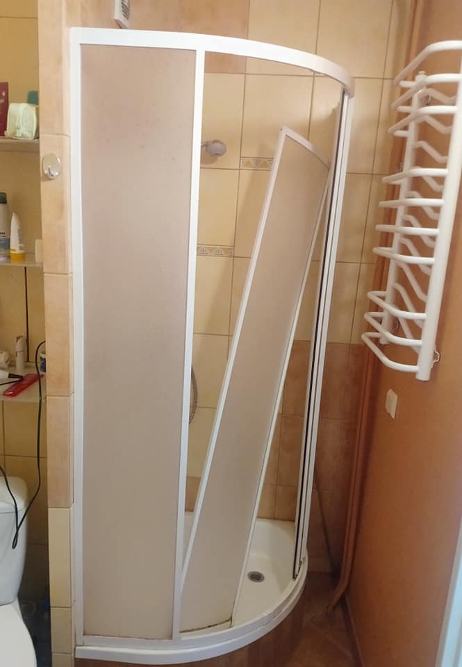 Narożna kabina prysznicowa z uchylnymi drzwiami w łazience z beżowymi płytkami, widoczny fragment toalety i grzejnik drabinkowy.